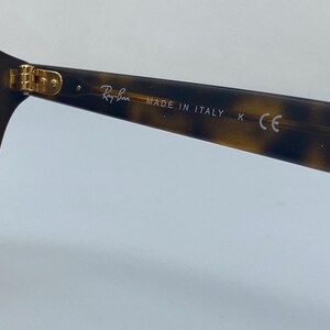 May include: Close-up of the tortoiseshell frame of a pair of Ray-Ban sunglasses. The frame has gold accents and the text "Ray-Ban MADE IN ITALY K CE" is printed on the side.