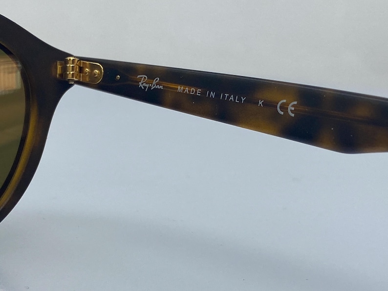 May include: Close-up of the tortoiseshell frame of a pair of Ray-Ban sunglasses. The frame has gold accents and the text "Ray-Ban MADE IN ITALY K CE" is printed on the side.