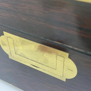 May include: Close-up of a wooden box with a dark brown finish and brass hardware. The brass hardware includes a rectangular plate with a handle-like design. The wood grain is visible.