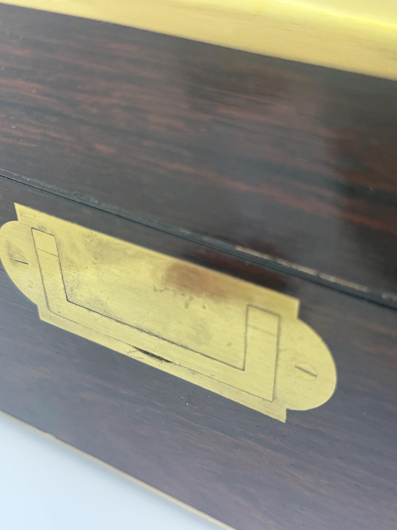 May include: Close-up of a wooden box with a dark brown finish and brass hardware. The brass hardware includes a rectangular plate with a handle-like design. The wood grain is visible.