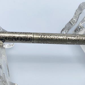 May include: Silver-toned pen with intricate floral engraving. The pen features a clip and a pointed tip. The design is detailed and the pen is displayed on a clear stand. A classic writing instrument.