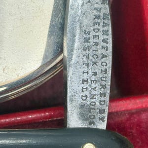May include: Close-up of a vintage straight razor with a black handle and a silver blade. The blade is inscribed with the text "Manufactured by Frederick Reynolds Sheffield". The razor is set against a red background.