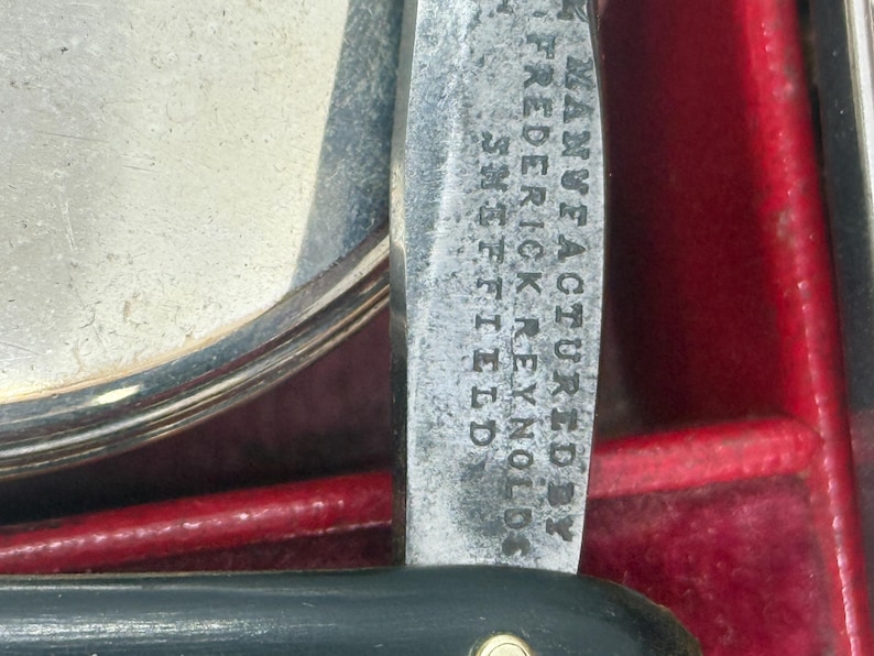 May include: Close-up of a vintage straight razor with a black handle and a silver blade. The blade is inscribed with the text "Manufactured by Frederick Reynolds Sheffield". The razor is set against a red background.