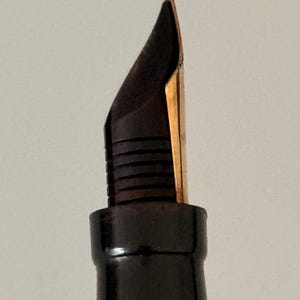 May include: A fountain pen nib made of gold-colored metal, featuring a pointed tip and a central slit. The nib is attached to a black plastic feed with horizontal ridges, which is connected to a smooth, black cylindrical pen grip section.