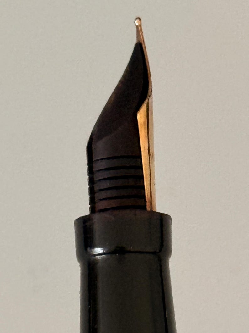 May include: A fountain pen nib made of gold-colored metal, featuring a pointed tip and a central slit. The nib is attached to a black plastic feed with horizontal ridges, which is connected to a smooth, black cylindrical pen grip section.