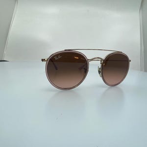 Puede incluir: Un par de gafas de sol redondas, de color oro rosa, con lentes marrones degradados. Las gafas tienen un fino marco de metal y un puente doble.