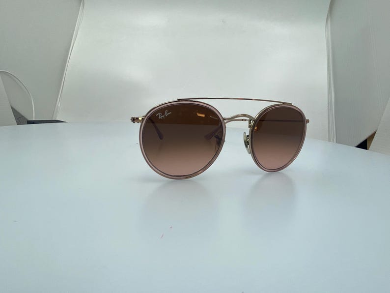 Puede incluir: Un par de gafas de sol redondas, de color oro rosa, con lentes marrones degradados. Las gafas tienen un fino marco de metal y un puente doble.