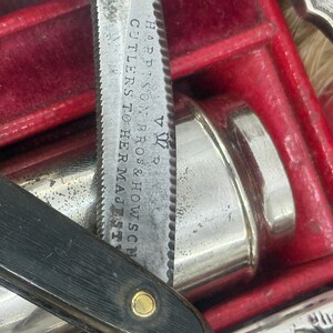 May include: Close-up of an antique straight razor with a black handle and a silver blade. The blade is marked with the text "HARRISON BROS & HOWSON CUTLERS TO HER MAJESTY". The razor is in a red velvet-lined case, alongside other silver items.
