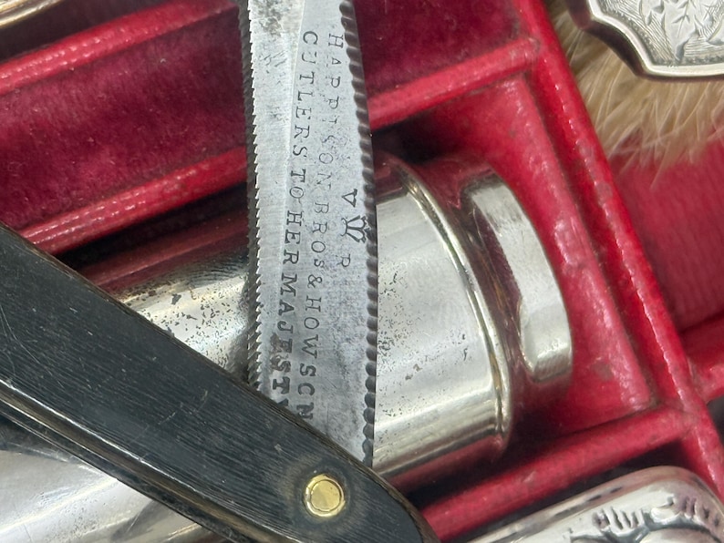 May include: Close-up of an antique straight razor with a black handle and a silver blade. The blade is marked with the text "HARRISON BROS & HOWSON CUTLERS TO HER MAJESTY". The razor is in a red velvet-lined case, alongside other silver items.