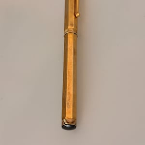 May include: A gold-toned hexagonal fountain pen featuring a metallic body with a matching cap and a pocket clip. The pen has a smooth, reflective finish and a dark, rounded end piece at the base, showcasing a sleek and elongated cylindrical design.