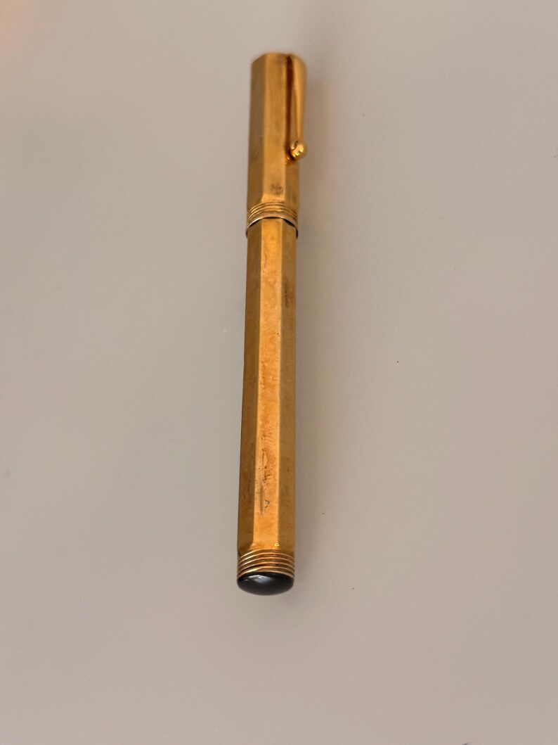 May include: A gold-toned hexagonal fountain pen featuring a metallic body with a matching cap and a pocket clip. The pen has a smooth, reflective finish and a dark, rounded end piece at the base, showcasing a sleek and elongated cylindrical design.