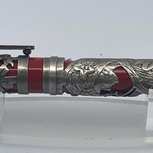 Peut inclure: Un stylo plume rouge doté d'un revêtement métallique argenté complexe avec des motifs de fleurs et de feuilles. Le stylo comprend un clip argenté assorti sur le capuchon et un corps rouge lisse, présentant un design en relief détaillé.