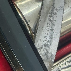 May include: Close-up of a vintage silver knife with a serrated edge. The blade is marked "Walter, Sheffield". The knife is partially visible, with a black handle and a red background.