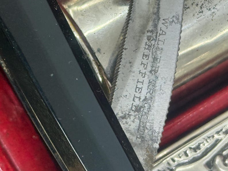 May include: Close-up of a vintage silver knife with a serrated edge. The blade is marked "Walter, Sheffield". The knife is partially visible, with a black handle and a red background.