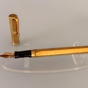 May include: A gold-toned fountain pen with a hexagonal barrel and a matching cap. The pen features a black grip section and a metallic nib. The cap includes a gold clip and is positioned upright next to the pen body, which rests horizontally.