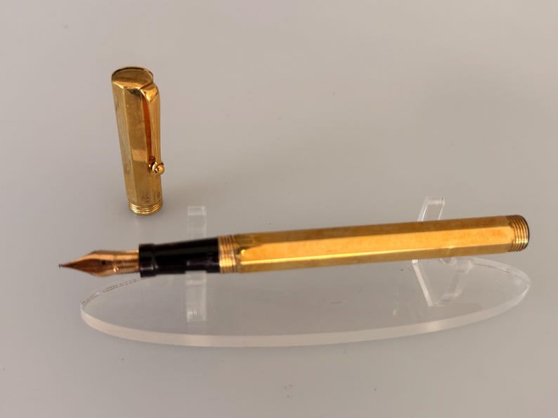 May include: A gold-toned fountain pen with a hexagonal barrel and a matching cap. The pen features a black grip section and a metallic nib. The cap includes a gold clip and is positioned upright next to the pen body, which rests horizontally.