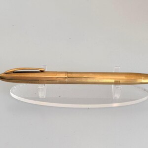 May include: A gold-colored fountain pen with a clip, resting on a clear acrylic stand. The pen has a smooth, metallic finish and a rounded tip. The pen is a classic writing instrument.