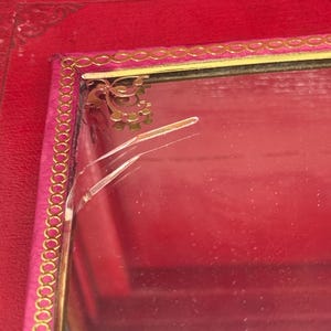May include: A red jewelry box with a gold trim and a pink interior. The box has a decorative gold design in the corner and a cracked glass surface. The box is made of a leather-like material.