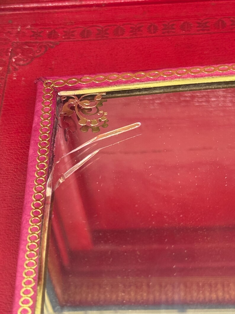 May include: A red jewelry box with a gold trim and a pink interior. The box has a decorative gold design in the corner and a cracked glass surface. The box is made of a leather-like material.