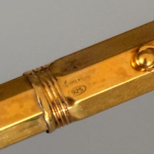 May include: A gold-toned metal bangle bracelet with a hexagonal profile and a ribbed decorative band. The surface features a stamped hallmark with the number 925 inside an oval, indicating sterling silver composition with a gold-colored finish.