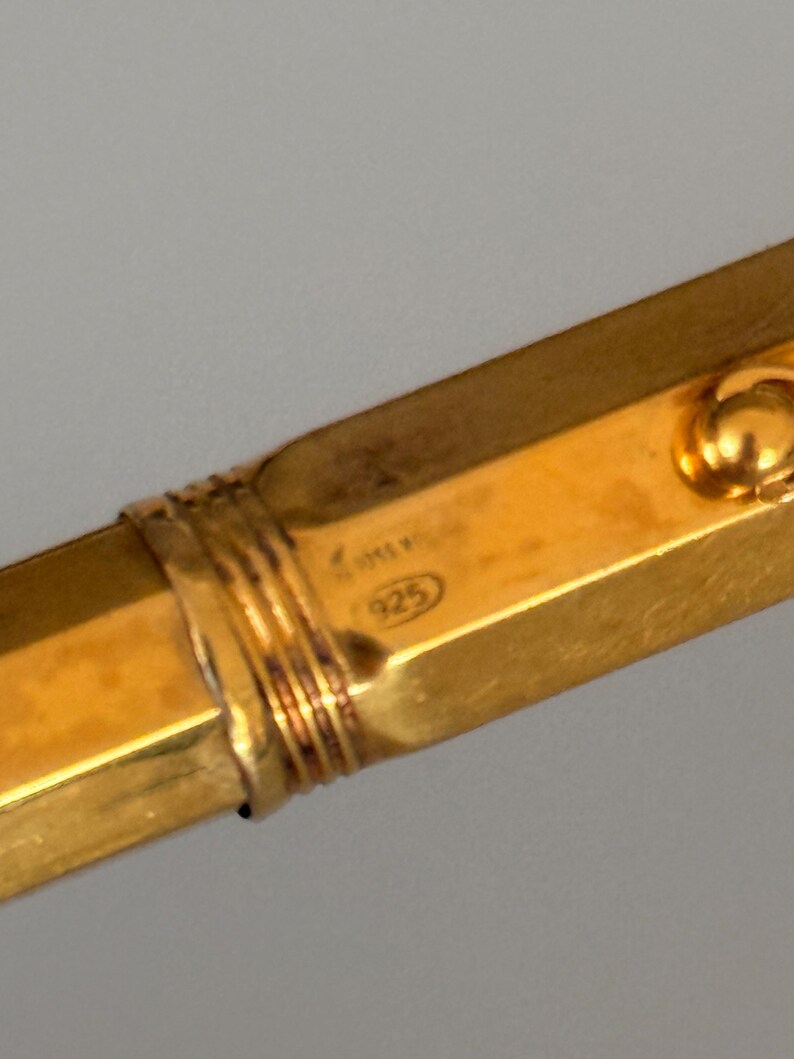 May include: A gold-toned metal bangle bracelet with a hexagonal profile and a ribbed decorative band. The surface features a stamped hallmark with the number 925 inside an oval, indicating sterling silver composition with a gold-colored finish.