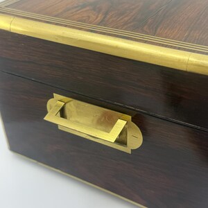 May include: A dark brown wooden box with brass accents. The box features a brass handle and trim around the edges. The top of the box has a floral brass inlay. The wood grain is visible.
