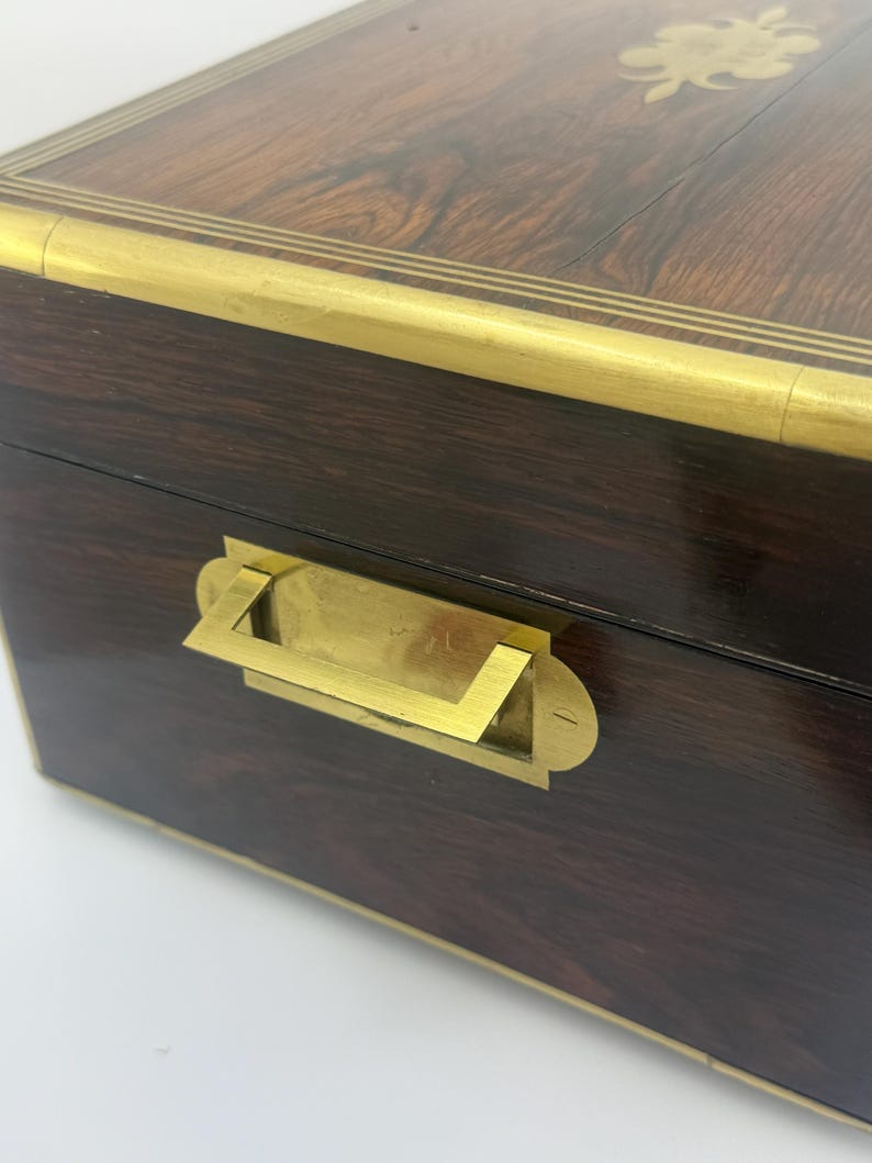 May include: A dark brown wooden box with brass accents. The box features a brass handle and trim around the edges. The top of the box has a floral brass inlay. The wood grain is visible.