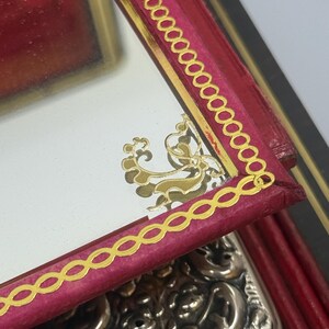 May include: Close-up of a red leather box with a gold-trimmed mirror. The mirror has a gold decorative design. The box has a gold chain-link border. A silver, ornate object is partially visible.