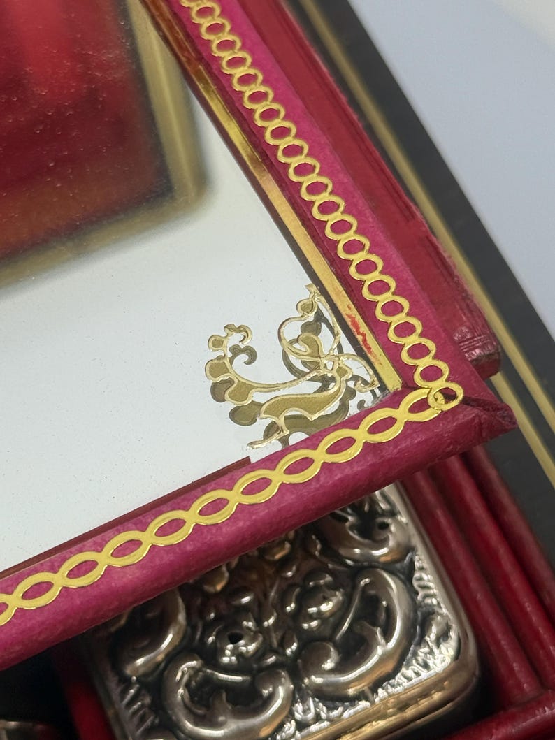 May include: Close-up of a red leather box with a gold-trimmed mirror. The mirror has a gold decorative design. The box has a gold chain-link border. A silver, ornate object is partially visible.