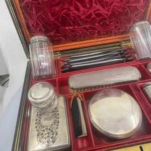 May include: An antique vanity set with a red velvet interior and various silver-toned grooming tools. The set includes jars, a brush, a compact, and other accessories, all neatly arranged within the black and gold-trimmed case. The case is open, revealing the contents.