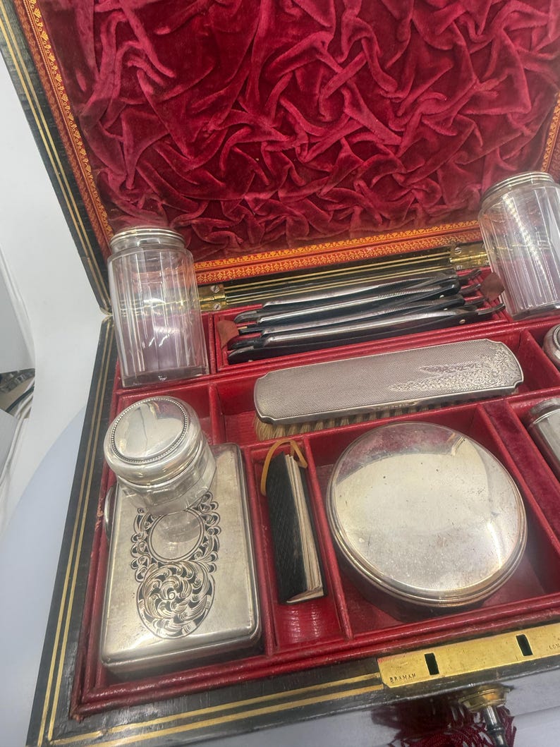May include: An antique vanity set with a red velvet interior and various silver-toned grooming tools. The set includes jars, a brush, a compact, and other accessories, all neatly arranged within the black and gold-trimmed case. The case is open, revealing the contents.