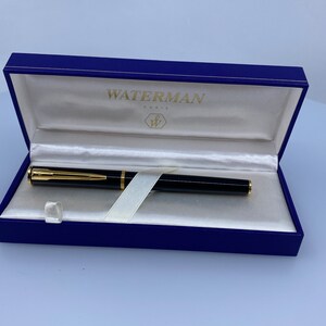 Pode incluir: Uma caneta-tinteiro Waterman preta e dourada numa caixa azul com o nome da marca "WATERMAN PARIS" na tampa.