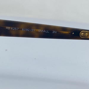 May include: Close-up of the inside of a tortoiseshell-colored pair of sunglasses with gold accents. The sunglasses have the text "RB 4257 6092/2Y SMALL 3N" printed on the inside of the frame.