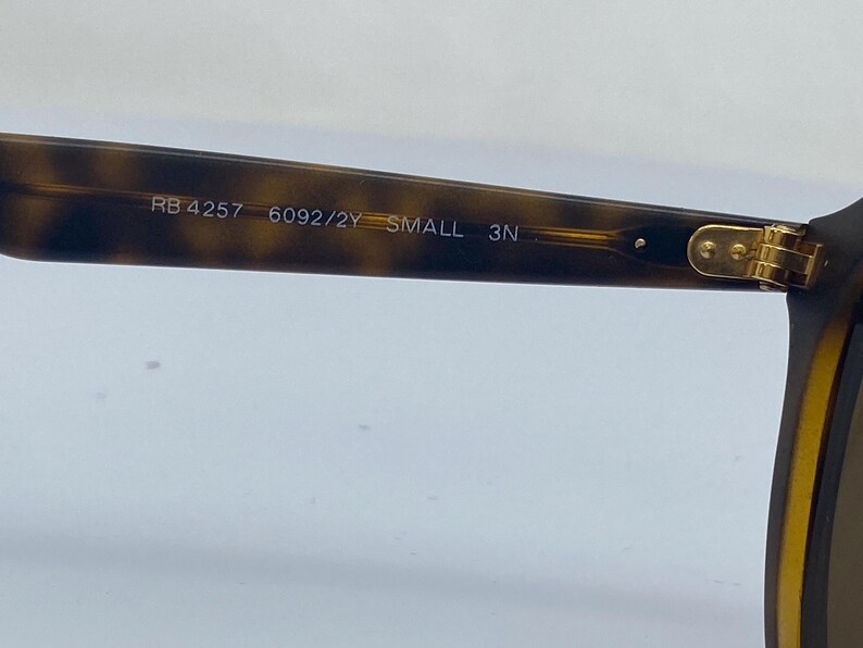 May include: Close-up of the inside of a tortoiseshell-colored pair of sunglasses with gold accents. The sunglasses have the text "RB 4257 6092/2Y SMALL 3N" printed on the inside of the frame.