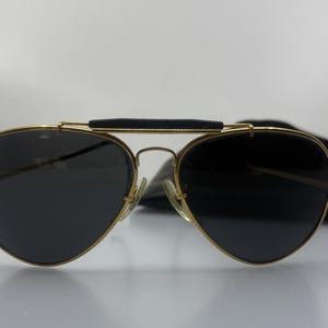 Könnte beinhalten: Goldfarbene Aviator-Sonnenbrille mit dunklen Gläsern. Die Sonnenbrille hat einen schwarzen Augenbrauensteg und ein schwarzes Etui ist im Hintergrund zu sehen. Die Gläser sind tropfenförmig.