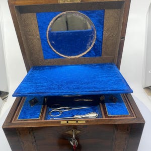 May include: An open, antique wooden box with a dark finish, inlaid with lighter wood accents. The interior is lined with blue velvet and features a small mirror and silver tools. The box is secured with a key.