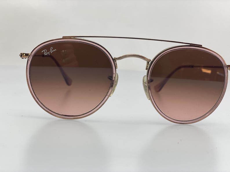 Puede incluir: Gafas de sol redondas con montura dorada y lentes de degradado rosa. Las gafas tienen una fina montura de metal y un logotipo de Ray-Ban en la lente izquierda.
