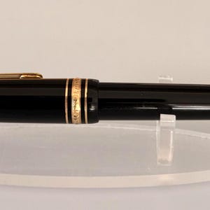 Könnte beinhalten: Ein schwarzer Kugelschreiber mit glänzender Oberfläche und goldfarbenen Akzenten am Clip, am Mittelring und an der spitz zulaufenden Spitze. Der Stift liegt horizontal auf einem ovalen, transparenten Acryl-Präsentationsständer.