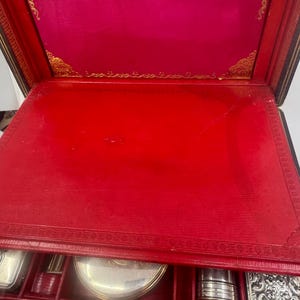 May include: An open, antique travel case with a red interior and gold accents. The case contains several silver-colored grooming accessories. The exterior is black with gold trim and a gold clasp. The interior lid is a deep pink color.