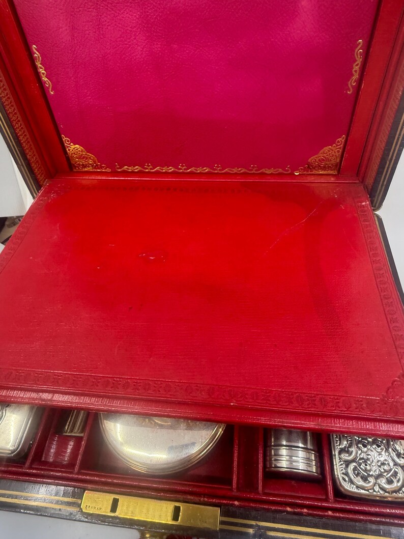 May include: An open, antique travel case with a red interior and gold accents. The case contains several silver-colored grooming accessories. The exterior is black with gold trim and a gold clasp. The interior lid is a deep pink color.