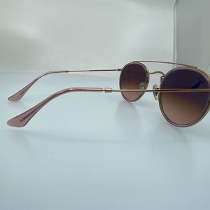 Puede incluir: Gafas de sol redondas de oro rosa con lentes marrones degradados. Las gafas tienen un fino marco de metal y se muestran de lado.