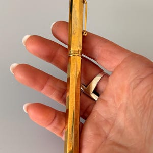 May include: A gold-toned fountain pen with a faceted, hexagonal barrel and cap. The pen features a slim, integrated pocket clip with a small spherical end. The surface of the metal shows a polished finish with subtle vertical texture and light wear.