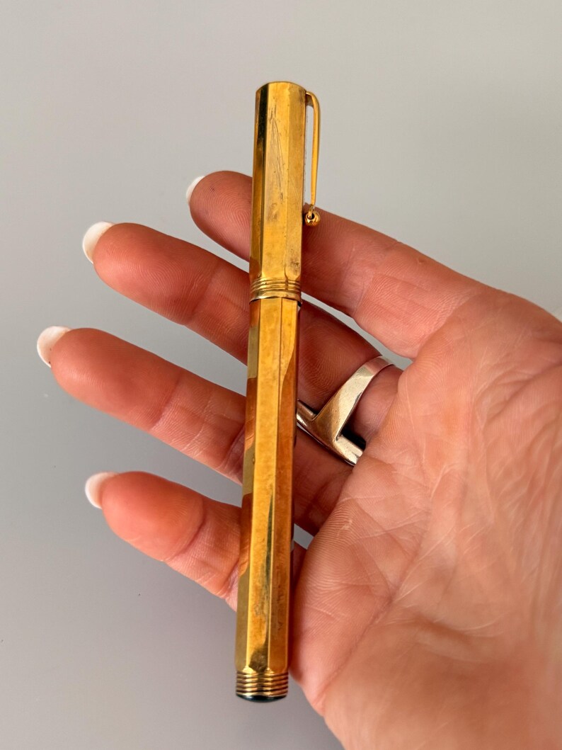May include: A gold-toned fountain pen with a faceted, hexagonal barrel and cap. The pen features a slim, integrated pocket clip with a small spherical end. The surface of the metal shows a polished finish with subtle vertical texture and light wear.