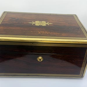 May include: A rectangular wooden box with a dark brown finish and gold-colored trim. The box has a keyhole and a decorative gold emblem on the lid. The wood grain is visible, and the box appears to be vintage.