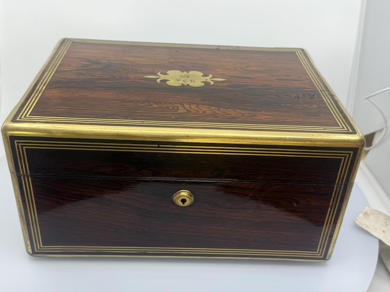 May include: A rectangular wooden box with a dark brown finish and gold-colored trim. The box has a keyhole and a decorative gold emblem on the lid. The wood grain is visible, and the box appears to be vintage.