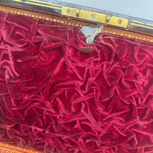 May include: An open antique box with a deep red velvet interior. The velvet is heavily textured and draped, filling the box. The box has a gold-colored trim and a metal clasp.