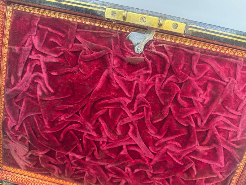 May include: An open antique box with a deep red velvet interior. The velvet is heavily textured and draped, filling the box. The box has a gold-colored trim and a metal clasp.