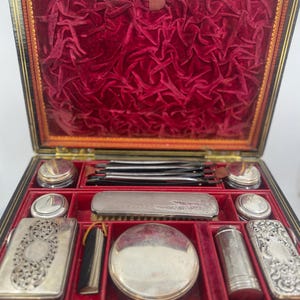 May include: An open antique vanity case with a black exterior and a red velvet interior. The case contains several silver-colored cosmetic and grooming tools, including compacts, jars, and a brush. The case is rectangular with a hinged lid.
