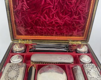 May include: An open antique vanity case with a black exterior and a red velvet interior. The case contains several silver-colored cosmetic and grooming tools, including compacts, jars, and a brush. The case is rectangular with a hinged lid.