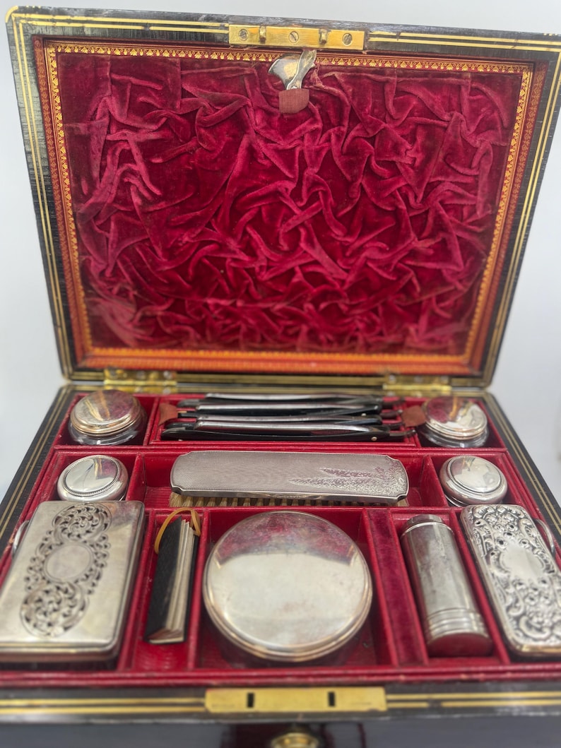 May include: An open antique vanity case with a black exterior and a red velvet interior. The case contains several silver-colored cosmetic and grooming tools, including compacts, jars, and a brush. The case is rectangular with a hinged lid.
